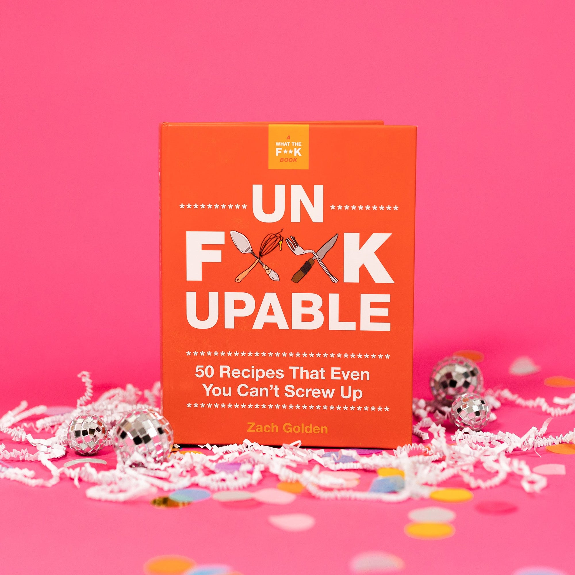 On a hot pink background sits a book with white crinkle and big, colorful confetti scattered around. There are mini disco balls. It is called "A WHAT THE F**K BOOK." It has illustraions of utensils crossed and in white it says "UN F**KUPABLE" in all caps, block font. It also says "50 Recipes that Even You Can't Screw Up" in white, block font. By Zach Golden.