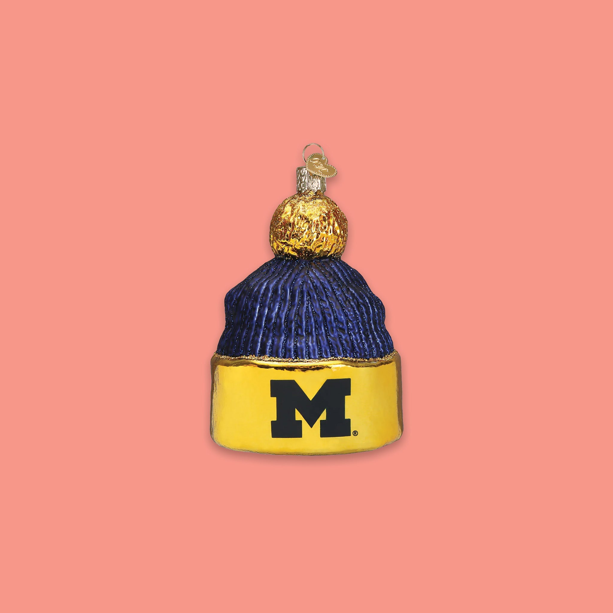 On a coral pink background sits a maize and navy University of Michigan beanie with a pom ball glass ornament. It has the letter "M" on it.