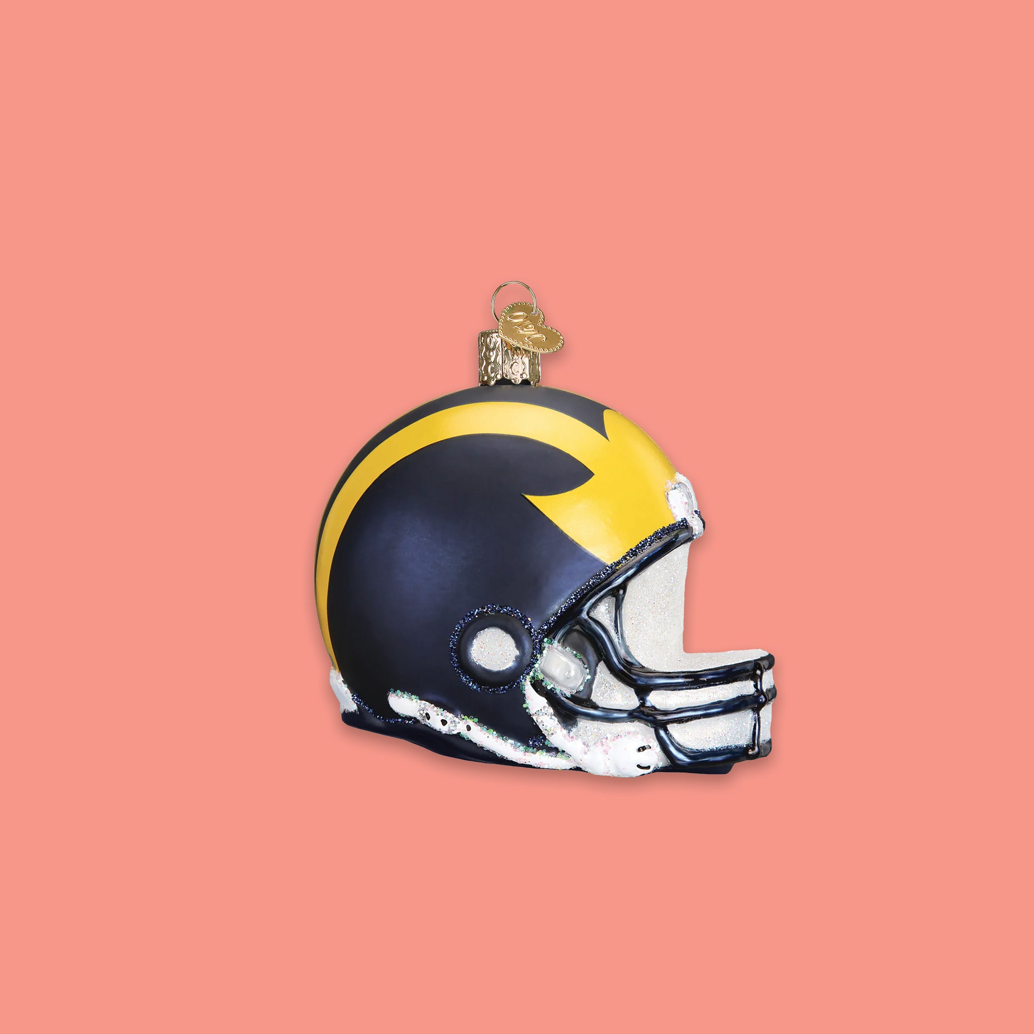 On a coral pink background sits a maize, navy, and white University of Michigan pennant glass ornament. It has the letter "M" on it.