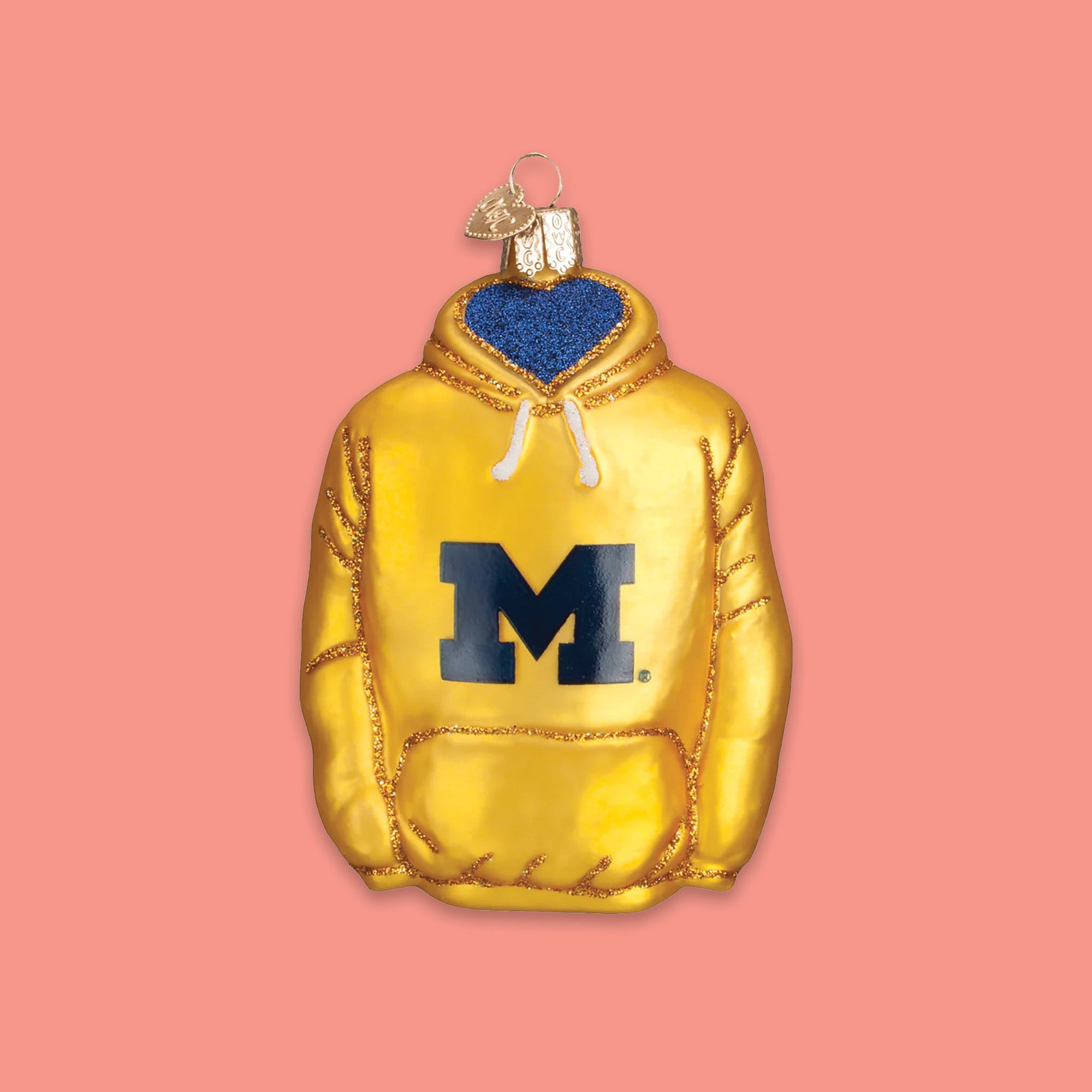 On a coral pink background sits a maize and navy University of Michigan hooded sweatshirt glass ornament. It has the letter "M" on it.