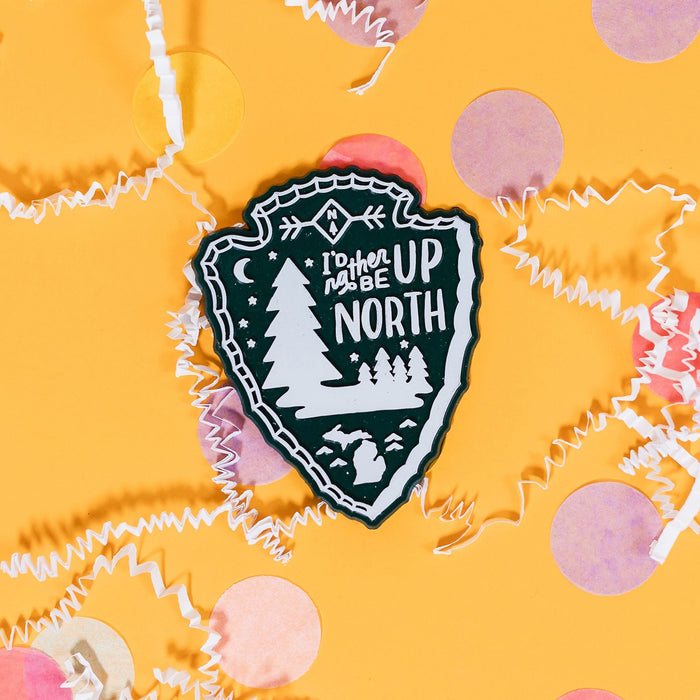 On a sunny mustard background sits a magnet with white crinkle and big, colorful confetti scatttered around. This 2D-style raised magnet is a dark green and white arrowhead with illustrations of white pine trees, half moon and state of Michigan and it says "I'd Rather Be Up North" in white handwritten lettering. 3"-4"