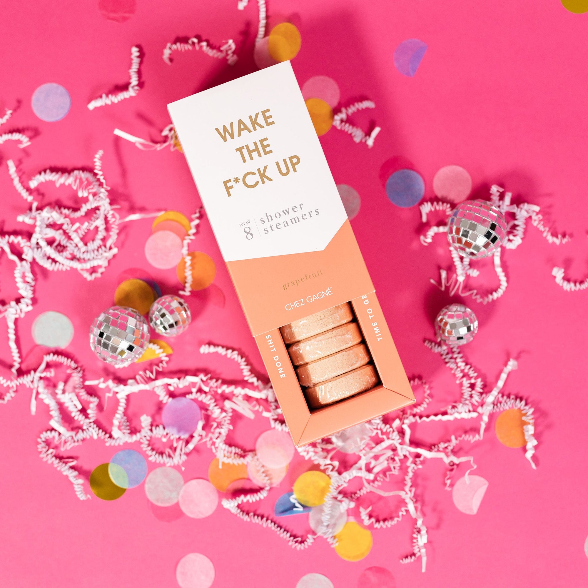 On a hot pink background sits an opened box with white crinkle and big, colorful confetti scattered around. There are mini disco balls. This picture is a close-up of a white and light pink package that says "WAKE THE F*CK UP" in gold foil, all caps block lettering. Under it says "set of 8" and " shower steamers" in grey, lowercase serif font. At the bottom it says "grapefruit" in gold foil, lower case serif font. The box is opened to reveal the peach shower steamers in it.