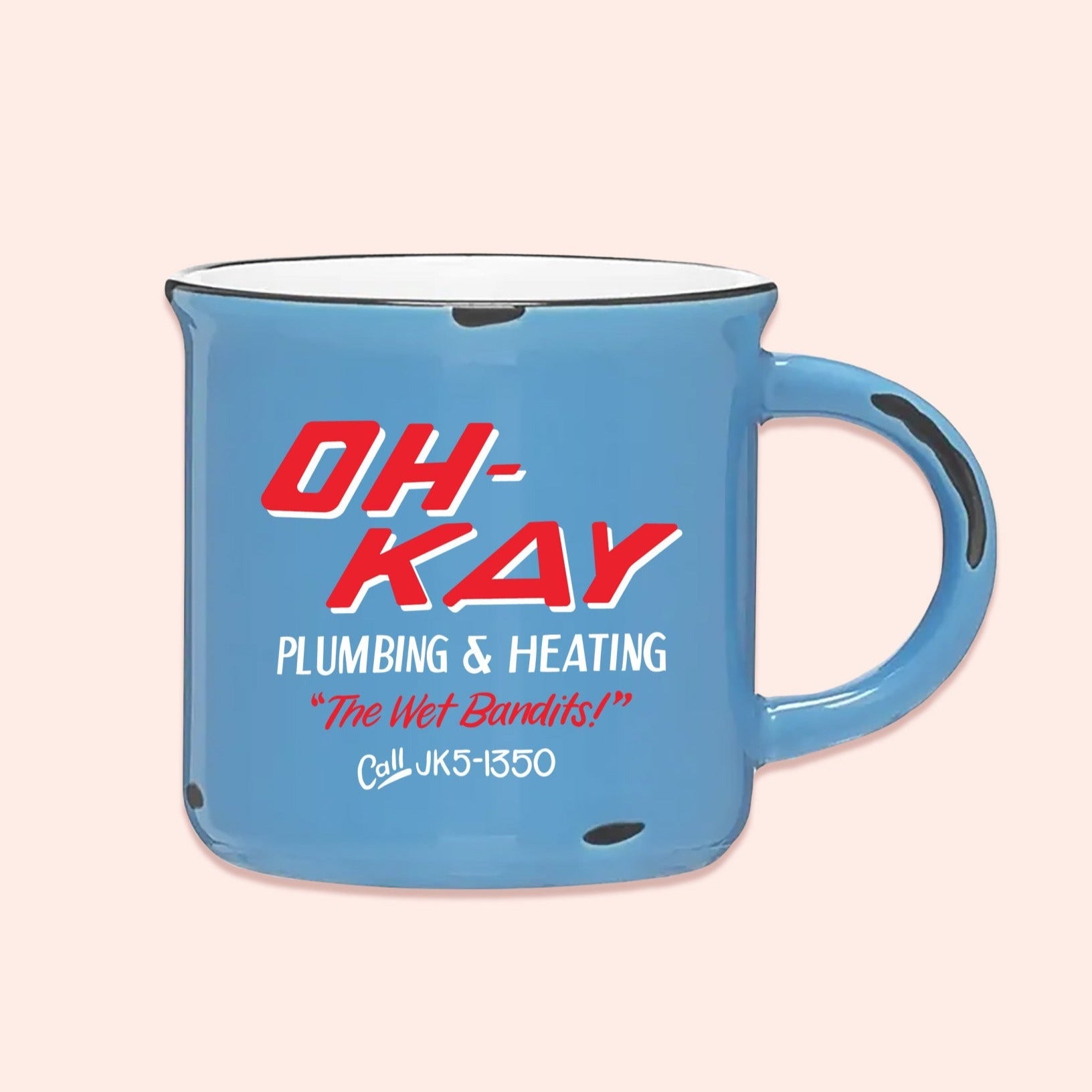 On a light pink background sits a campfire mug. This Home Alone inspired mug is light blue with white and bright coral red lettering. It says "OH-KAY PLUMBING & HEATING, "The Wet Bandits!", and Call JK5-1350."