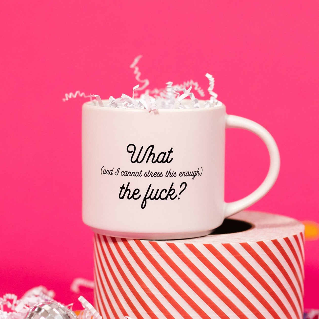 On a hot pink background sits a mug with white crinkle and big, colorful confetti scattered around. This white mug says "What (and I cannot stress this enough) the fuck?" in black script font. It has white crinkle in it and sits atop a red and white striped roll of tape. 