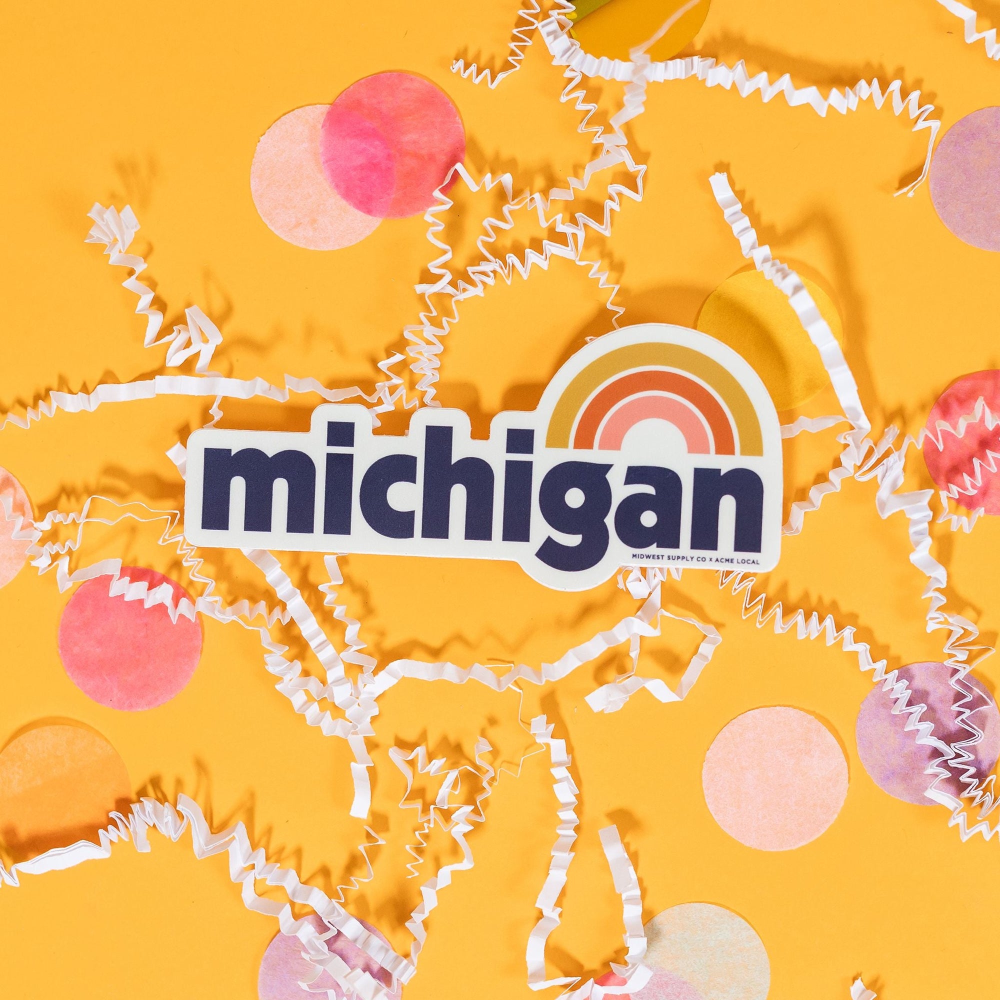 On a sunny mustard background sits a sticker with white crinkle and big, colorful confetti scattered around. This white sticker says "michigan" in a navy bold font with a rainbow above it in mustard yellow, orange, and peach. 2"-3"