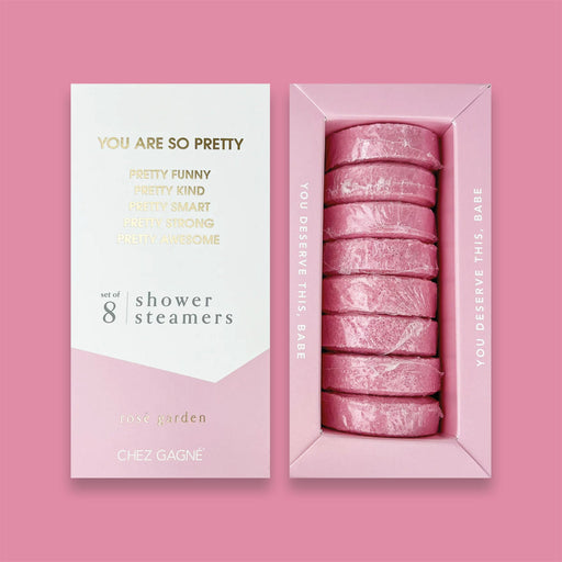 On a bubblegum pink background sits two boxes. This picture is a close-up of a white and light pink package that says "YOU ARE SO PRETTY" and "PRETTY FUNNY PRETTY KIND PRETTY SMART PRETTY STRONG PRETTY AWESOME" in gold foil, all caps block lettering. Under it says "set of 8" and " shower steamers" in grey, lowercase serif font. At the bottom it says "rosé garden" in gold foil, lower case serif font. To the right of it is a light pink box with pink shower steamers in it. 