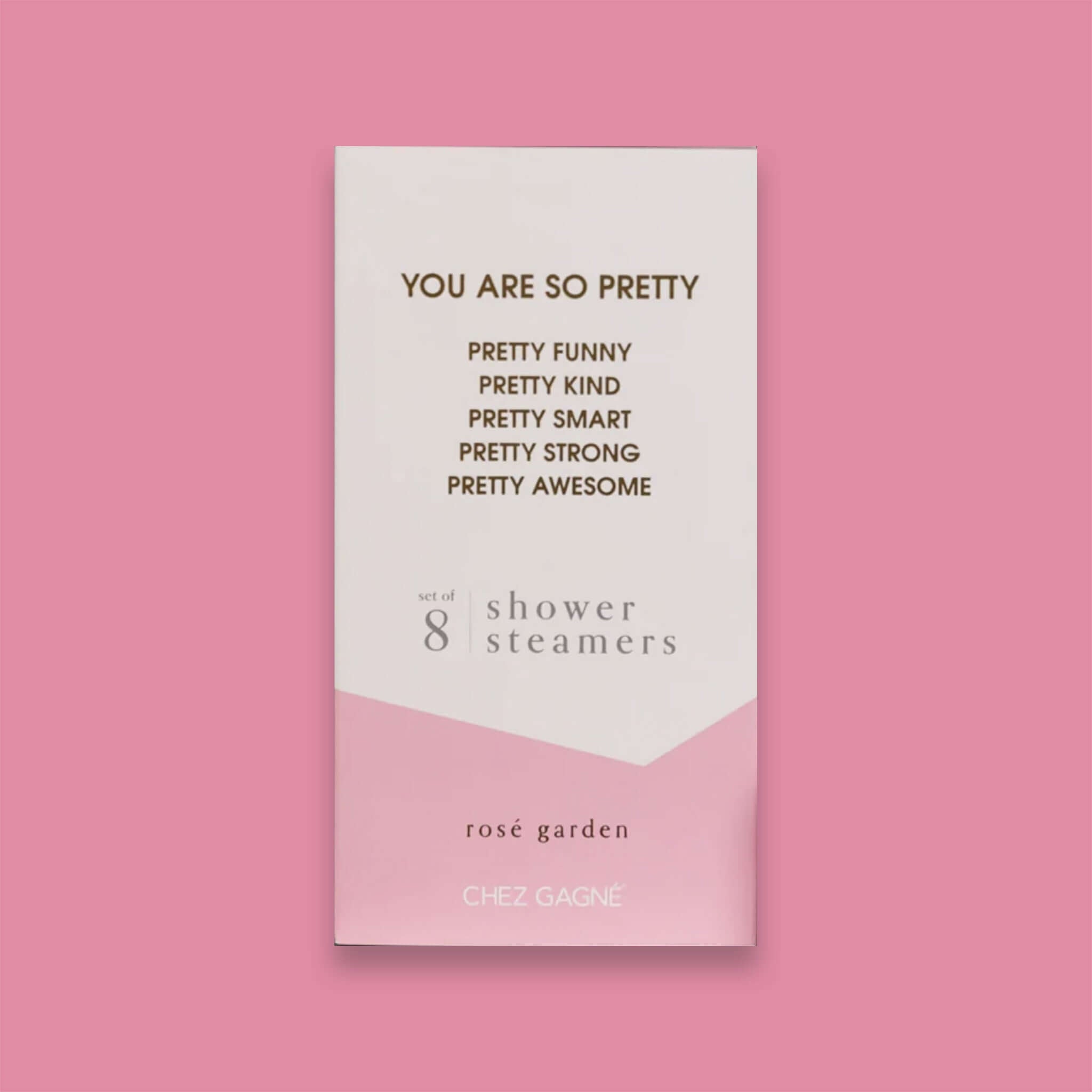 On a bubblegum pink background sits a box. This picture is a close-up of a white and light pink package that says "YOU ARE SO PRETTY" and "PRETTY FUNNY PRETTY KIND PRETTY SMART PRETTY STRONG PRETTY AWESOME" in gold foil, all caps block lettering. Under it says "set of 8" and " shower steamers" in grey, lowercase serif font. At the bottom it says "rosé garden" in gold foil, lower case serif font.