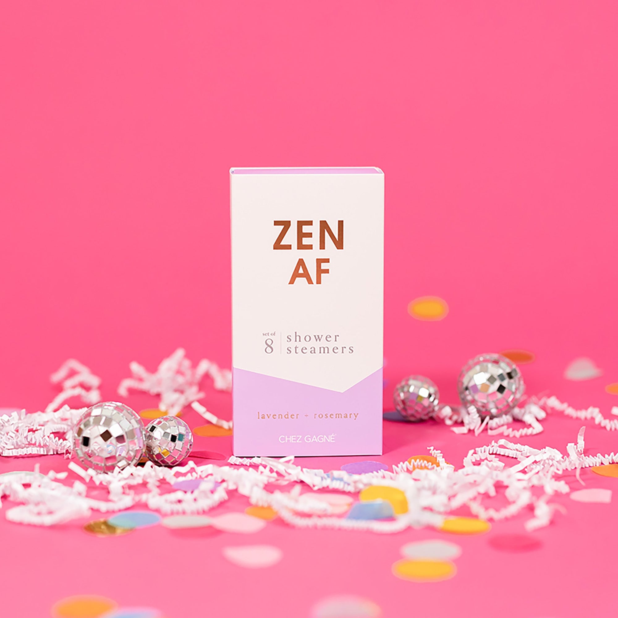 On a hot pink background sits a box with white crinkle and big, colorful confetti scattered around. There are mini disco balls. This picture is a close-up of a white and lavender package that says "ZEN AF" in gold foil, all caps block lettering. Under it says "set of 8" and " shower steamers" in grey, lowercase serif font. At the bottom it says "lavender - rosemary" in gold foil, lower case serif font. To the right of it is a lilac box with lavender shower steamers in it. 