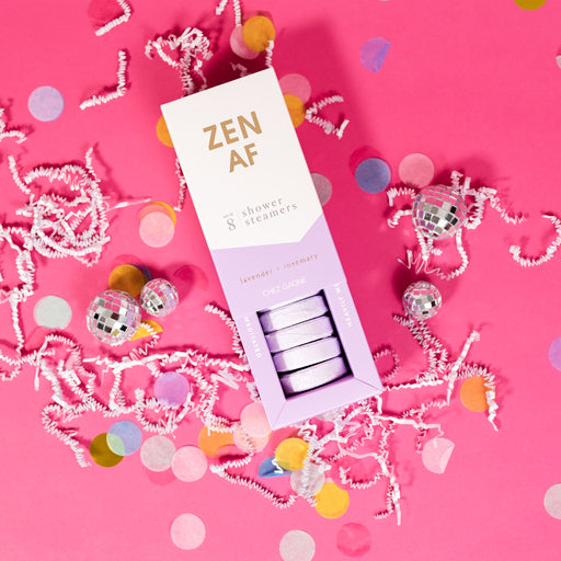 On a hot pink background sits an opened box with white crinkle and big, colorful confetti scattered around. There are mini disco balls. This picture is a close-up of a white and lavender package that says "ZEN AF" in gold foil, all caps block lettering. Under it says "set of 8" and " shower steamers" in grey, lowercase serif font. At the bottom it says "lavender - rosemary" in gold foil, lower case serif font. The box is opened to reveal the lavender shower steamers in it.