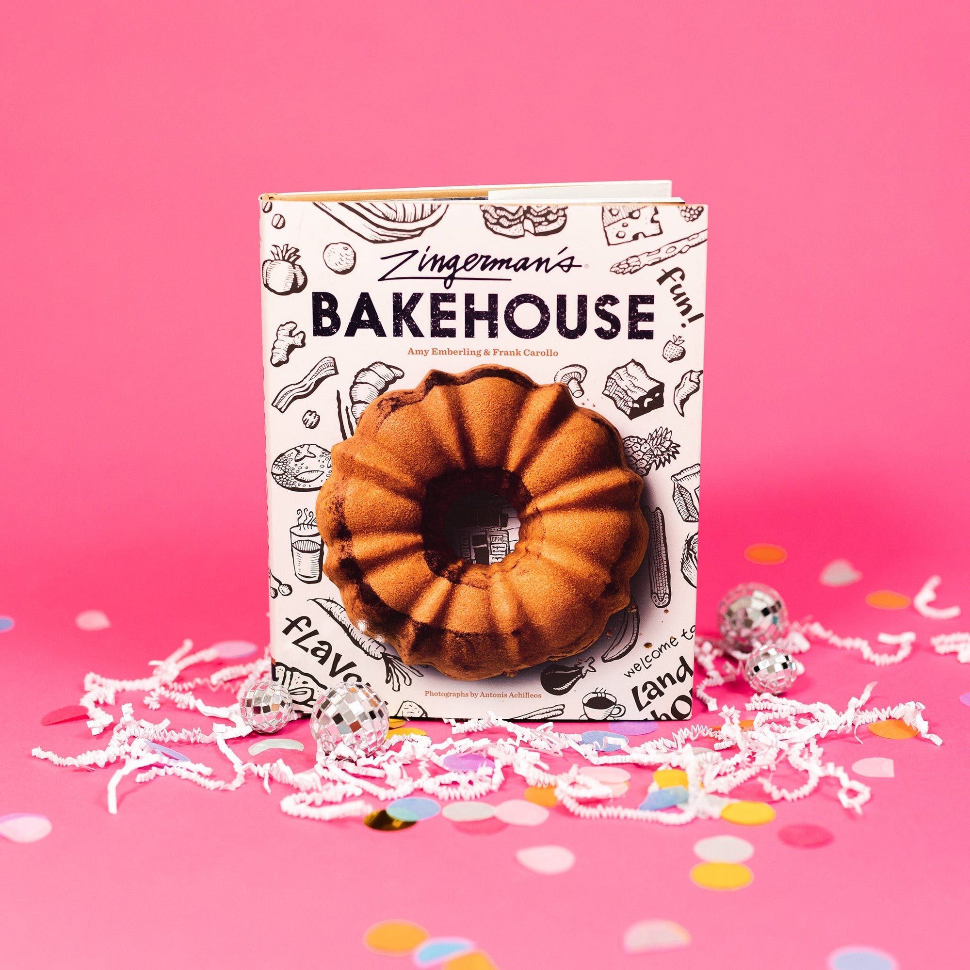 On a hot pink background sits a book with white crinkle and big, colorful confetti scattered around. There are mini disco balls. This cookbook says "Zingerman's Bakehouse" in a dark purple font. The cover has black illustrations of fruit, vegetables, bacon, drinks and a photo of a bundt cake.