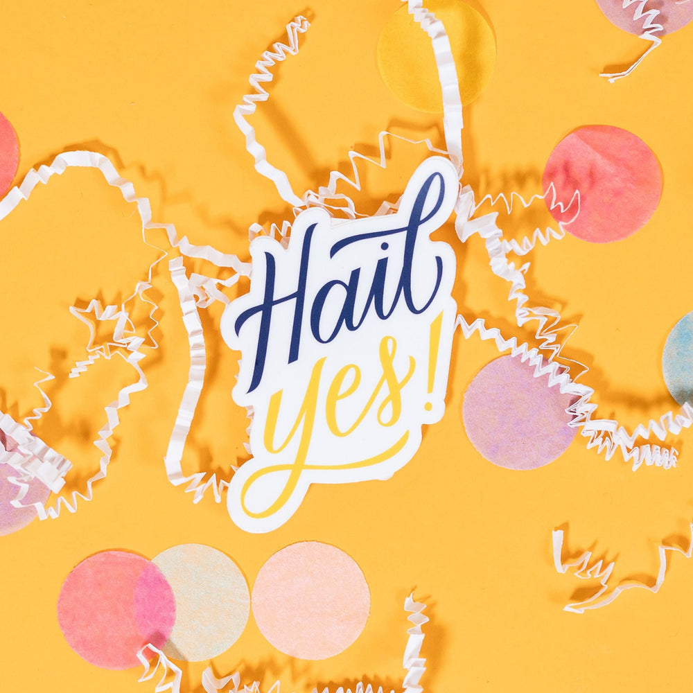 On a sunny mustard background sits a sticker with white crinkle and big, colorful confetti scattered around. This white sticker says "Hail Yes!" in handdrawn script lettering. The "Hail" is in navy and the "Yes!" is in mustard yellow. 2"-3"