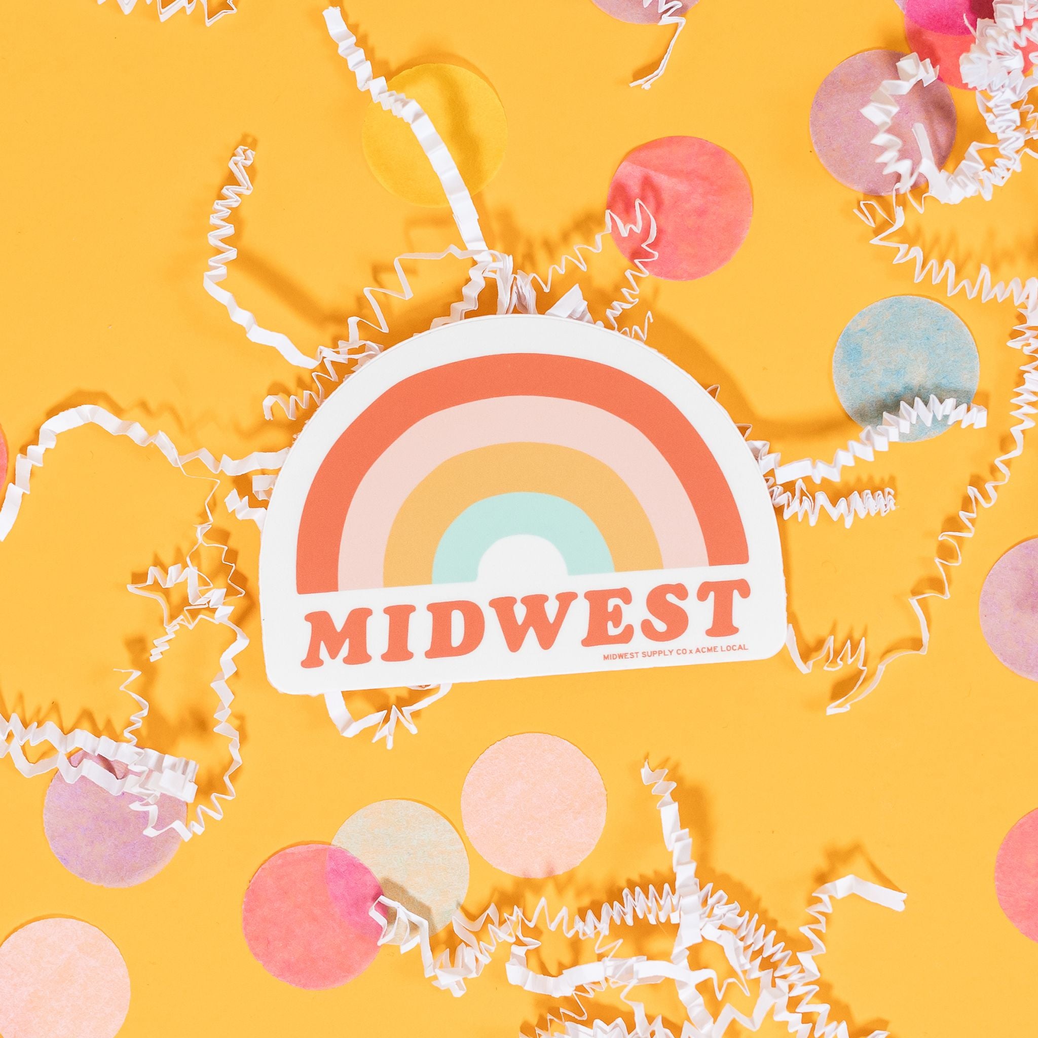 On a sunny mustard background sits a sticker with white crinkle and big, colorful confetti scattered around. This white sticker has an illustration of a rainbow in orange, pink, mustard yellow, and aqua blue. It says "MIDWEST" in an orange, fat serif font. 2"-3"