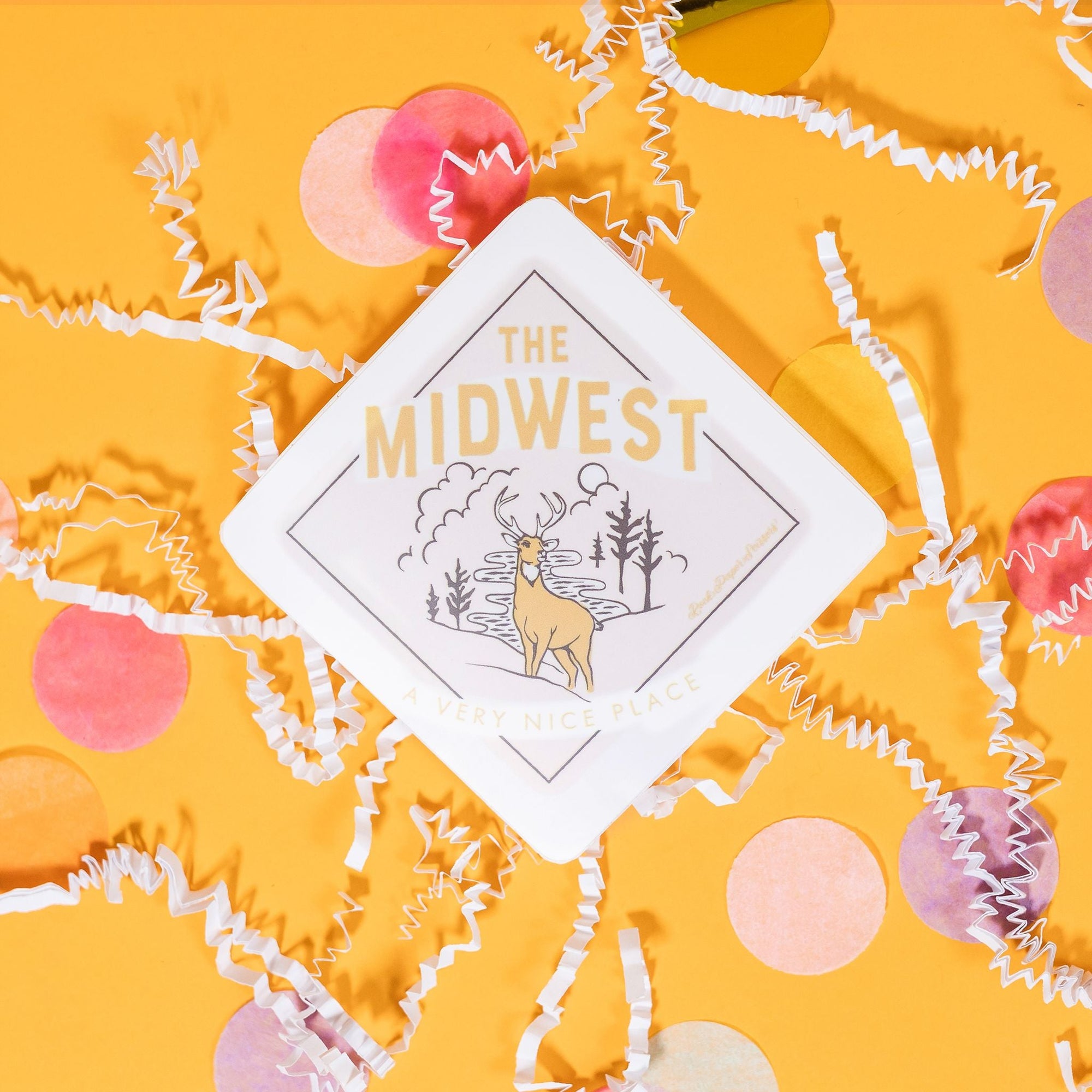 On a sunny mustard background sits a sticker with white crinkle and big, colorful confetti scattered around. This white, diamond sticker has an illustration of a stag deer with pine trees, clouds, sun, and river. It is in a light orange yellow with brown. It says "THE MIDWEST", "A Very Nice Place"" in light orange yellow block lettering. 2"-3"