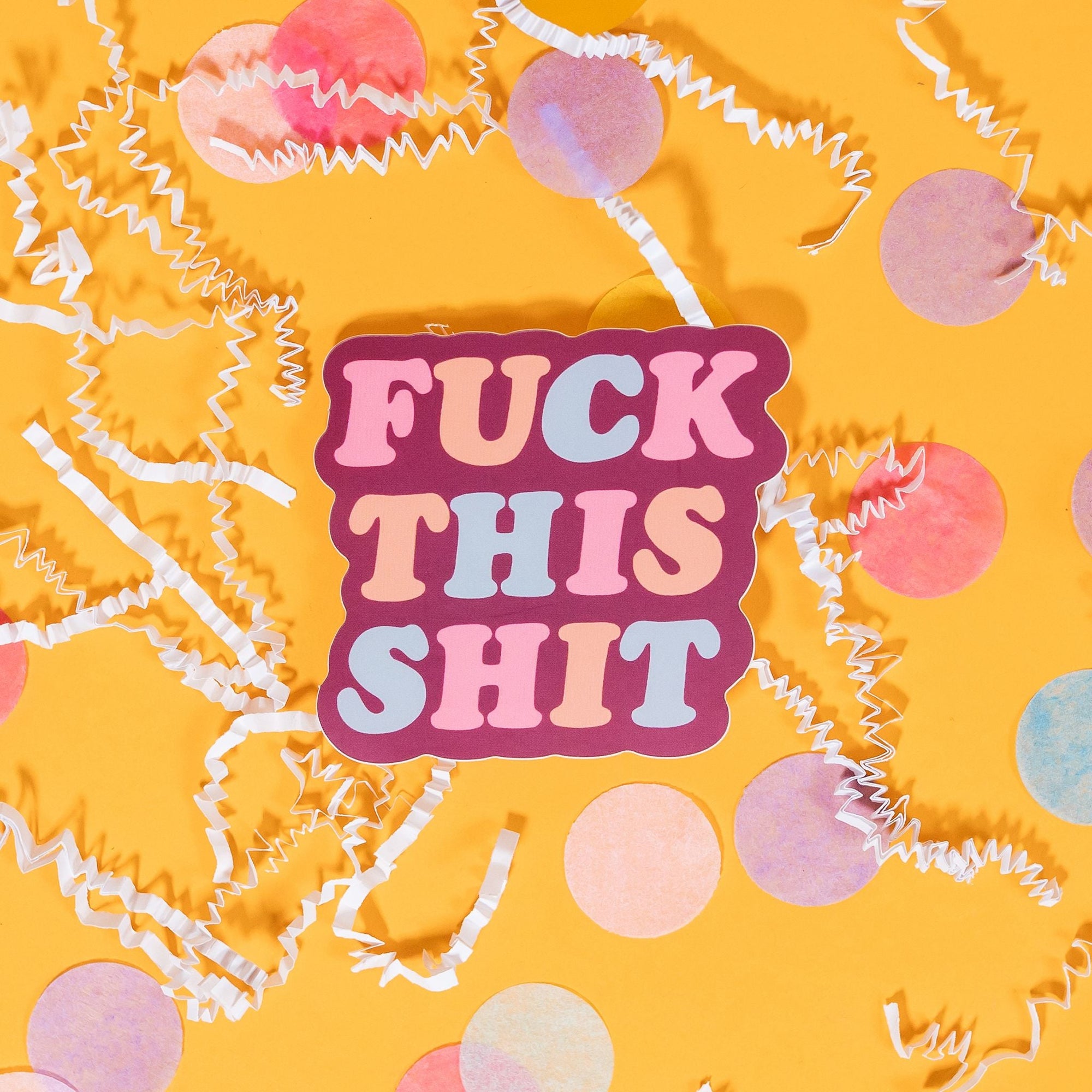 On a sunny mustard background sits a sticker with white crinke and big, colorful confetti scattered around. This raspberry colored sticker says "FUCK THIS SHIT" in a fat, serif font in pink, peach and baby blue.