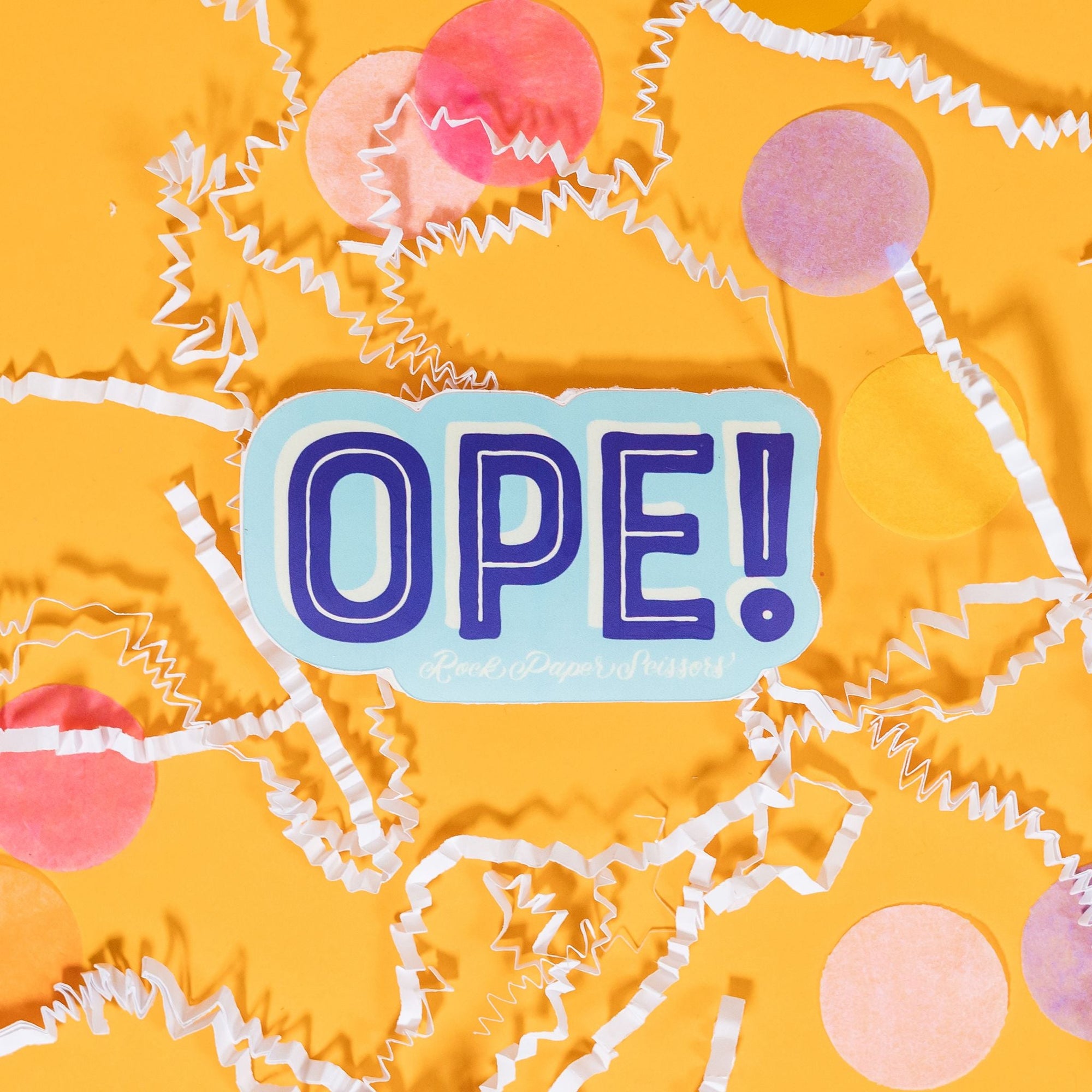 On a sunny mustard background sits a sticker with white crinkle and big, colorful confetti scattered around. This sky blue sticker says "OPE!" in handwritten brilliant blue block lettering and white skinny handwritten lettering on top, with a white dropshadow. Under it says "Rock Paper Scissors" in white handwritten script lettering. 2"-3"