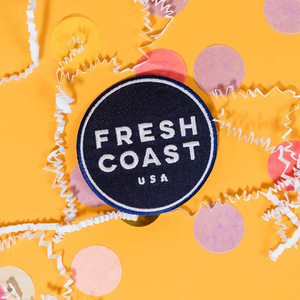 On a sunny mustard background sits a patch with white crinkle and big, colorful confetti scattered around. This navy patch has a white outline circle stitching on the edge and in the center in white it says "FRESH COAST USA" in block stitch lettering.