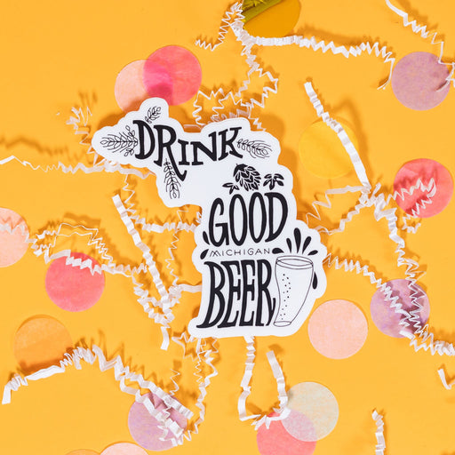 On a sunny mustard background sits a sticker with white crinkle and big, colorful confetti scattered around. This white sticker has a handdrawn illustration of a beer glass with hops in black and it says "Drink Good Michigan Beer" in black handwritten lettering. 2"-3"
