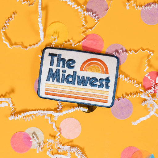 On a sunny mustard background sits a patch with white crinkle and big, colorful confetti scattered around. This white patch has navy outline stitching on the edge and in the center in white it says "The Midwest" in navy block stitch lettering. There is a rainbow on top in orange, coral and mustard yellow stitching and on the bottom are three lines in orange, coral and mustard yellow stitching.