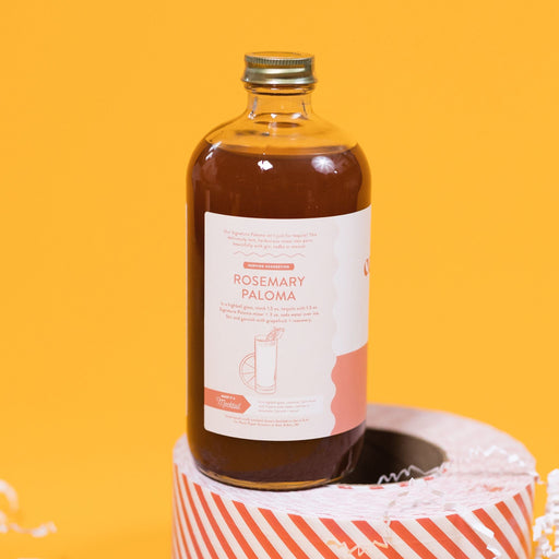 On a sunny mustard background sits a bottle and packing tape surrounded by white crinkle. This close-up of the clear bottle is a Rock Paper Scissors Cocktail Co. Drink Mixer called "Signature Paloma". The flavor is rosemary grapefruit. It has a white label with a grapefruit pink stripe at the bottom. The bottle sits atop a red and white striped packing tape. 16 oz (473ml)