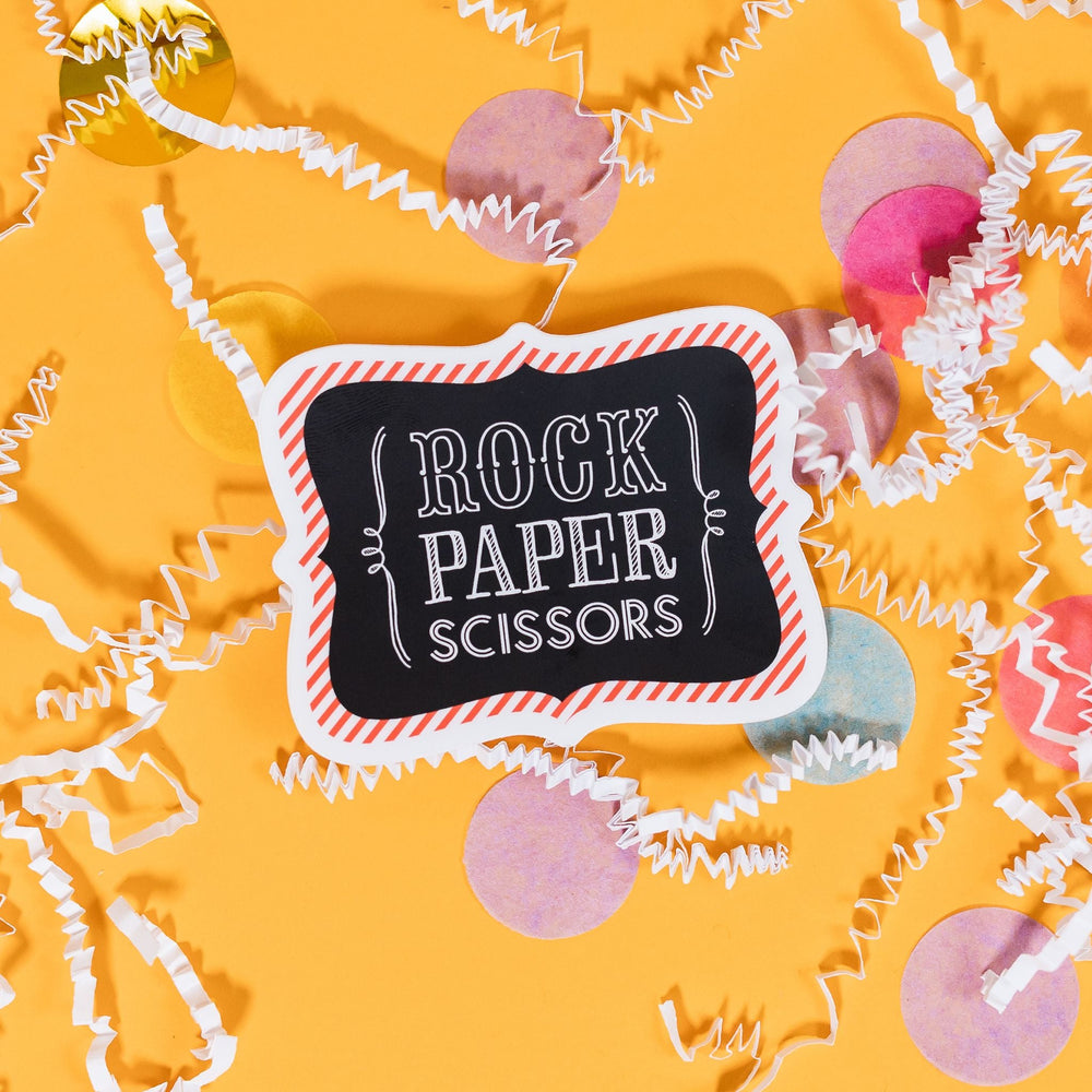 On a sunny mustard background sits a sticker with white crinkle and big, colorful confetti scattered around. The cartouche shaped sticker is white with a red and white striped border. In the middle it is black and on top of the black it says "Rock Paper Scissors" in white handwritten lettering. 2"-3"
