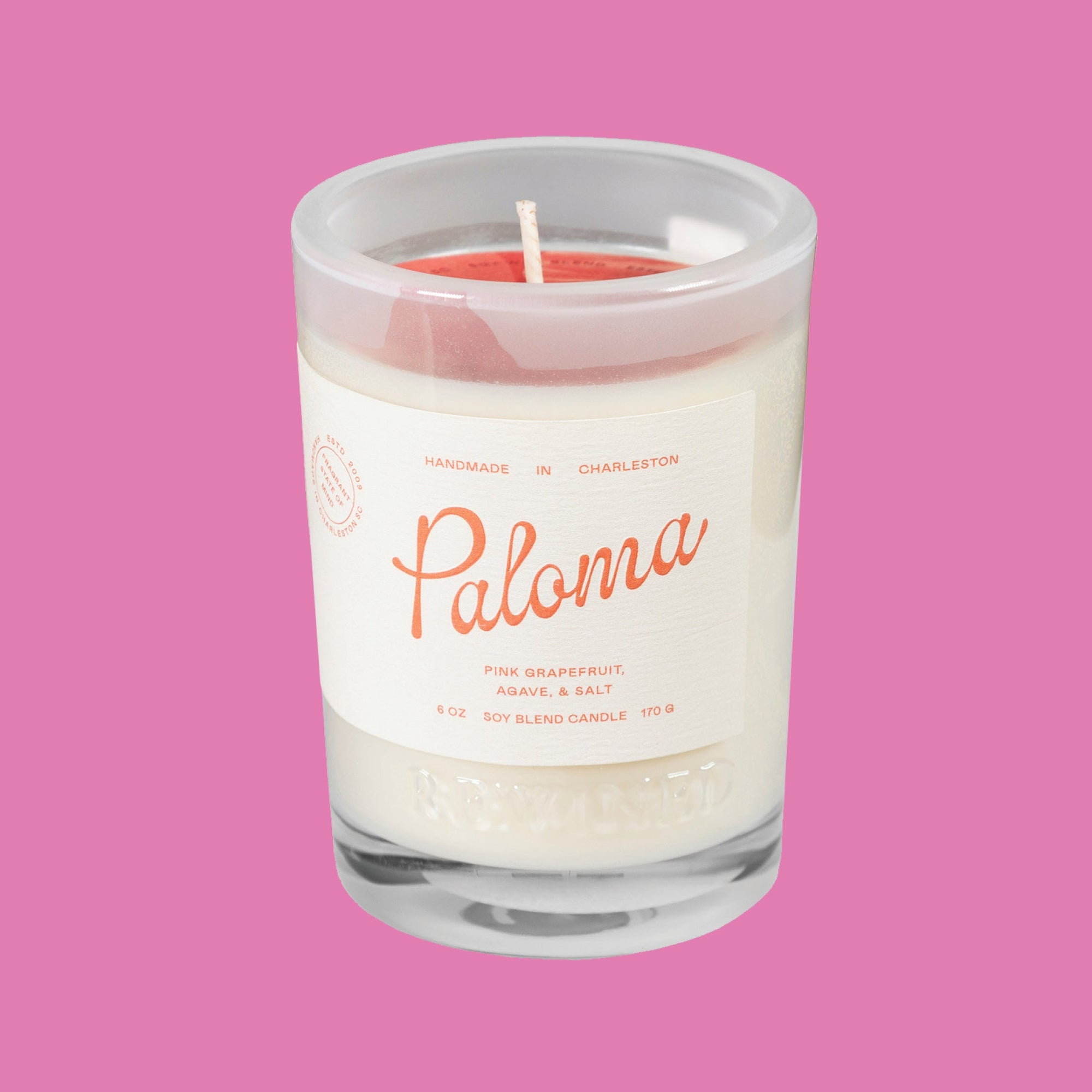 On a raspberry pink background sits a Rewined Sparkling Collection Handmade Paloma Soy Blend Candle. It's scent is pink grapefruit, agave, and salt. It has a cream label with coral text.